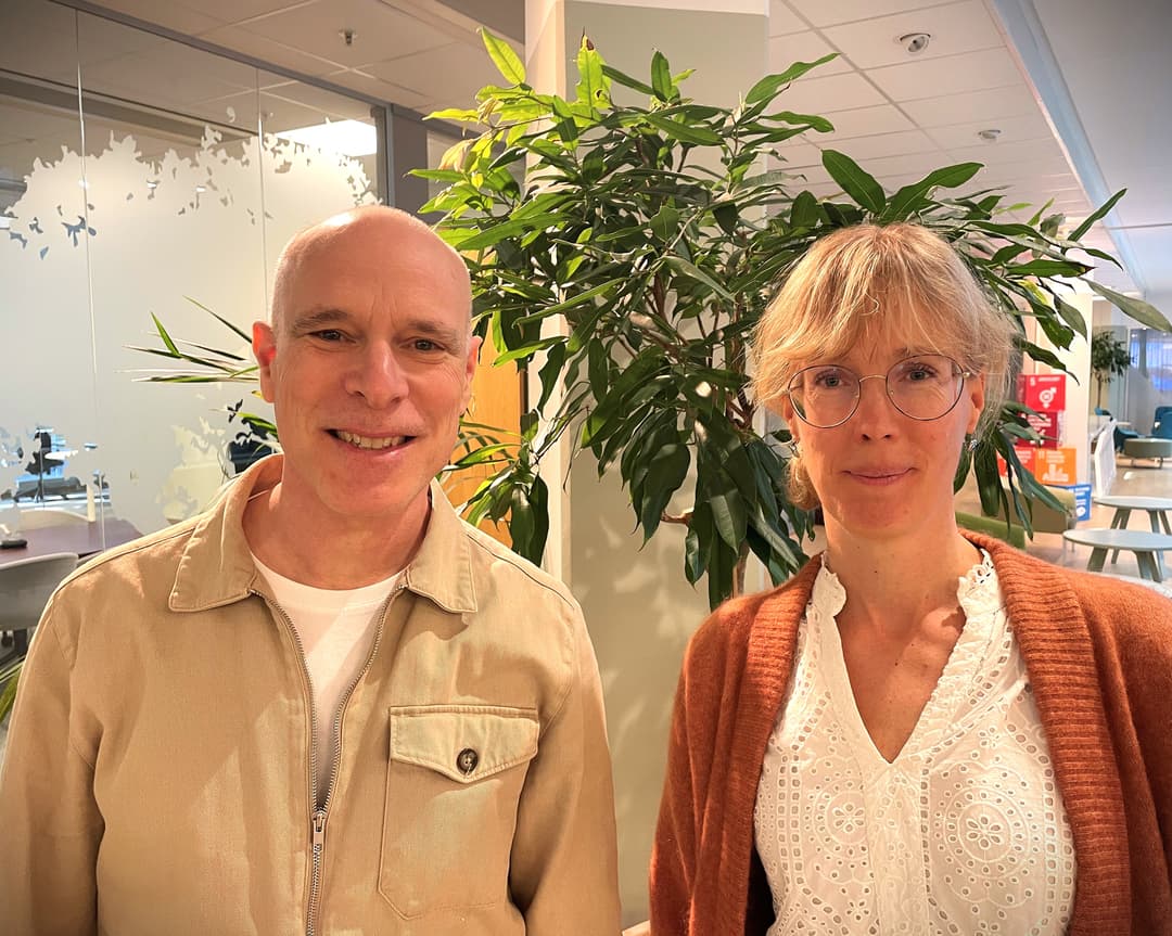 En bild på en man och kvinna från Folkhälsomyndigheten. Mannen står till vänster och kvinnan till höger. I bakgrunden finns et kontorslandskap och gröna växter