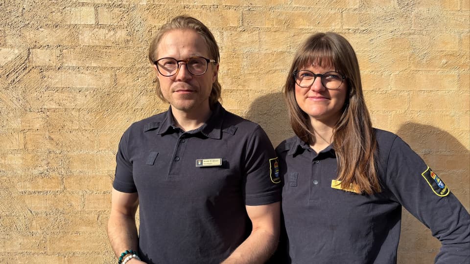Två personer står framför en beige husfasad iklädda uniformer från Kriminalvården